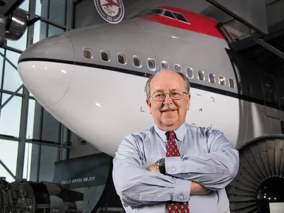 A white man in his early 70s stands inside a museum gallery. He is wearing glasses and smiling. Behind him, is the nose and forward fuselage of a Boeing 747 airplane. The 747 is painted in a red, gray, and white paint scheme.