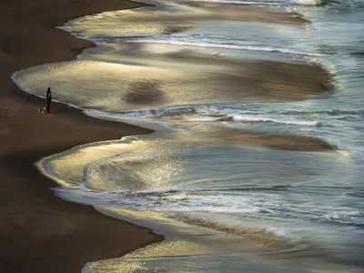 At dawn, a woman and her dog walk along a serene shore in the Corona Del Mar area of Newport Beach as gentle waves greet them.