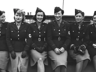 Six women sitting closely together posing for the camera. They are wearing uniforms with light skirts and dark jackets and an armband with the letter C on it.