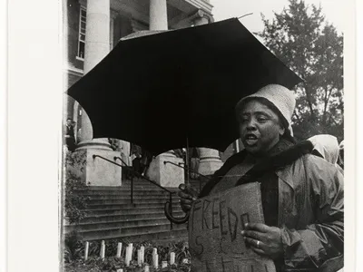 A black-and-white photograph of Fannie Lou Hamer carrying a sign and holding an umbrella over her head.