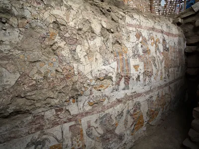 A wall painting of a crowned woman with scepter (upper left), a procession of men behind her carrying objects (upper right) and a textile workshop (below)