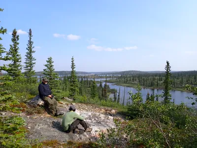 Two men sit on a rock bed on top of a forested hill over looking a remote lake.